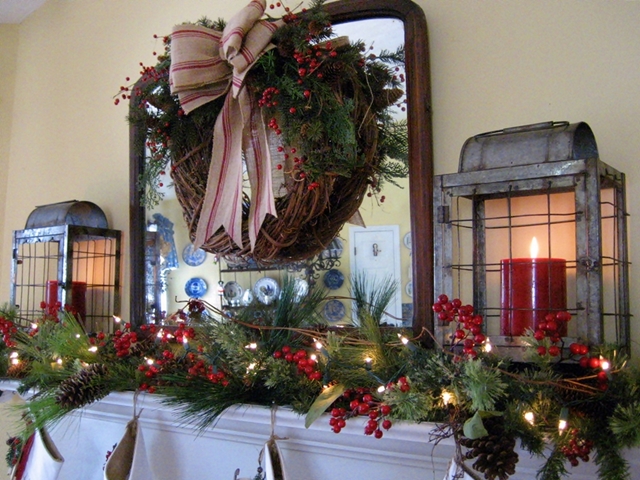 my farmhouse christmas mantel, christmas decorations, seasonal holiday decor, wreaths, Lots of greenery with red berries rustic lanterns and a grapevine wreath with a grain sack ribbon make up my farmhouse mantel