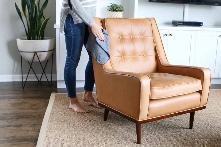 how to clean leather furniture, woman wearing jeans and a gray sweater wipes down a brown leather tufted chair with a gray towel