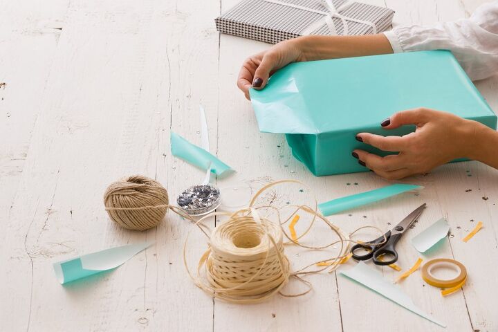 how to wrap a present, hands wrapping a gift in teal wrapping paper with more materials laid out on a white table