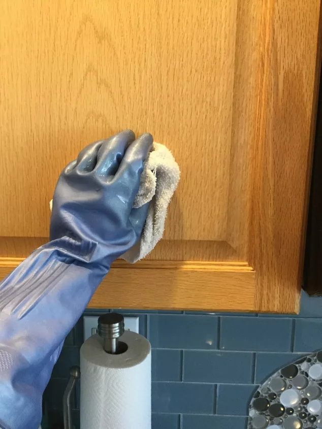 how to clean kitchen cabinets, rubber gloved hand wiping down wood kitchen cabinet door with a towel