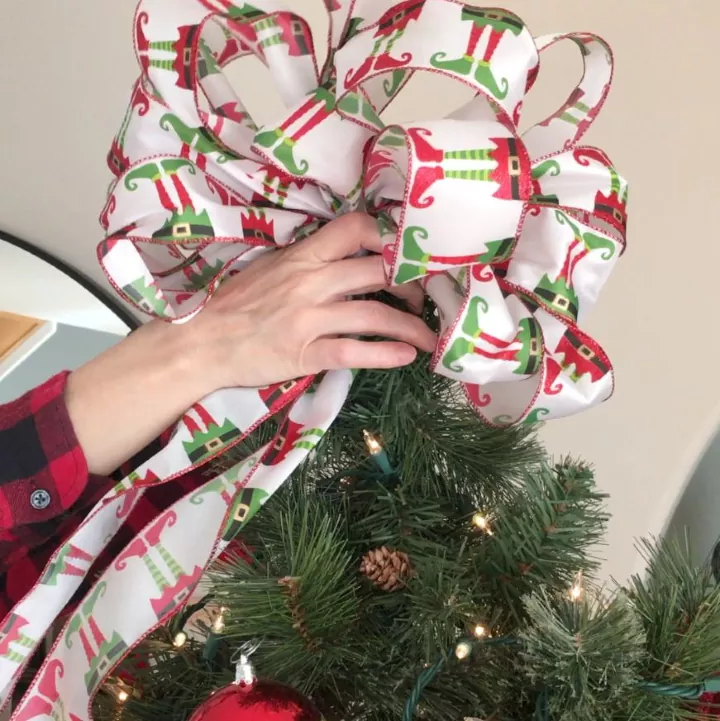 how to make a christmas bow, hand putting large elf printed Christmas bow on top of a tree