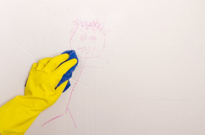 como limpar paredes de tinta plana e se livrar de marcas teimosas, M o amarela com luva de borracha usando um pano azul limpando um giz de cera da parede branca