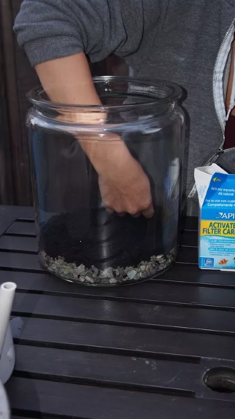 how to make a terrarium in 5 simple steps, hand putting pebbles and soil into glass container