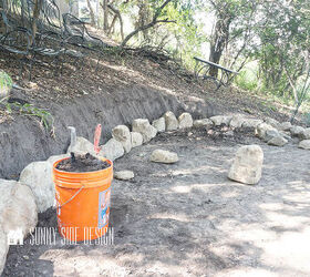 A orange bucket is filled with backfill dirt to backfill the first row of stones for the retaining wall