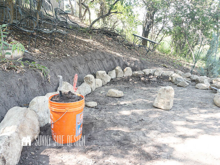 A orange bucket is filled with backfill dirt to backfill the first row of stones for the retaining wall