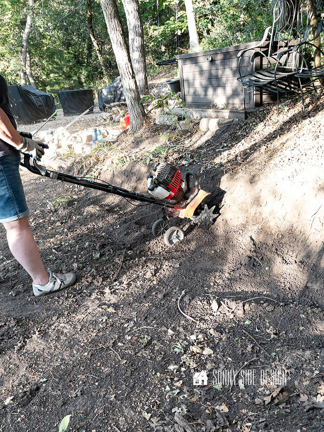 Woman uses a small garden tiller to loosen the hard clay soil to prepare the area for building a stone retaining wall
