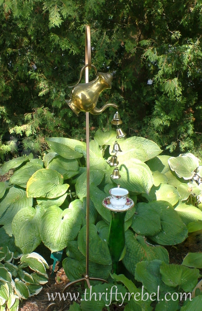 vintage coffee pot wind chimes, gardening, repurposing upcycling, Vintage Coffee Pot Wind Chimes Small Christmas Bells are tied to fishing line and are poured from the coffee pot spout into a lovely cup and saucer held up by a green bottle covered stake